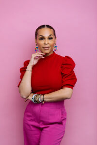 Mavic Bright, Mindset Coach, regarde avec détermination et confiance, vêtue d'un haut rouge vif et d'un pantalon fuchsia. Cheveux tirés en chignon bas, elle porte des boucles d'oreilles colorées et des bracelets à la main gauche. En arrière-plan, un rose bonbon vibrant. Une représentation puissante de l'estime de soi et de la confiance personnelle.